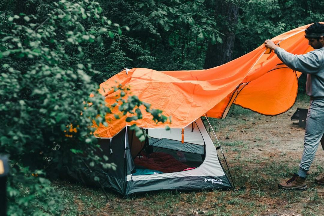 A camper stretches an orange rain fly over a gray tent at a wooded campsite